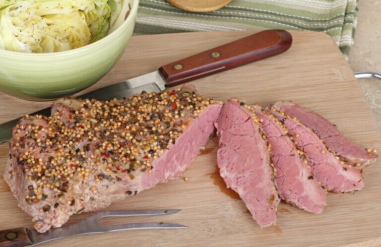 A fan of seasoned corned beef slices are shown sliced off of a larger steak, and displayed on a bamboo cutting board.