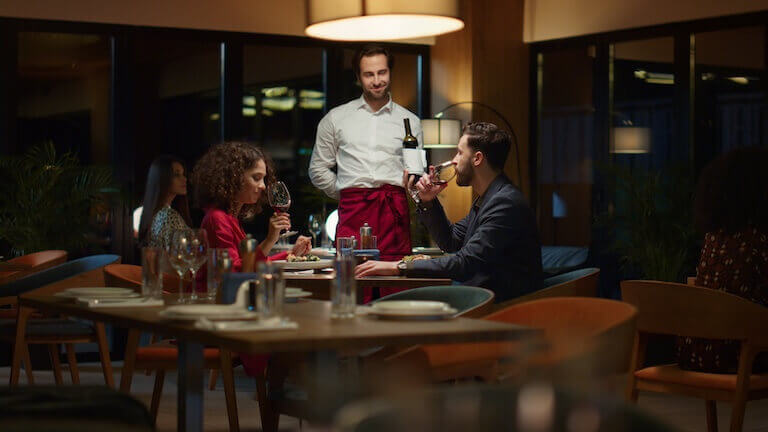 A server stands with a bottle of wine and looks on as a couple samples a glass of red wine in dimly lit fancy restaurant dining room.