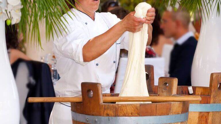 Cheesemaker making cheese in a wooden barrel as people dine in the background. 
