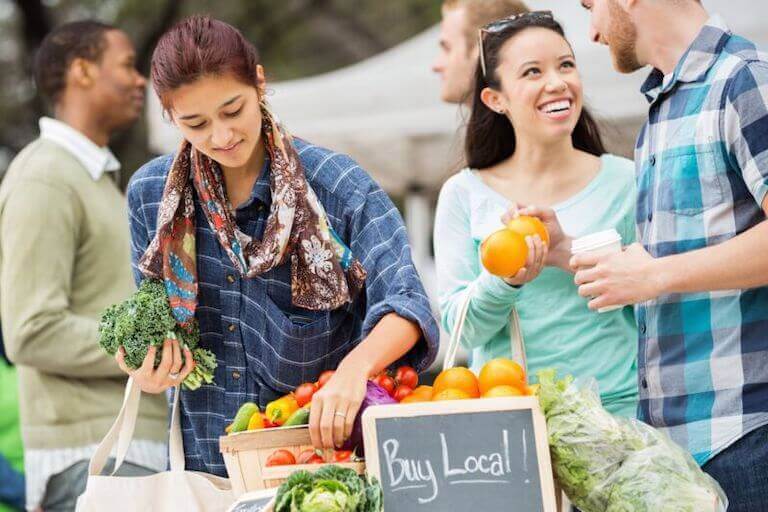 Customers sort through baskets of peppers, greens, and oranges at a farmer’s market near a chalkboard sign that says, “Buy Local!”