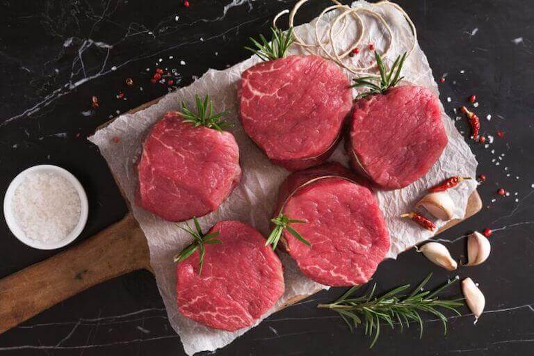 Raw filet mignon resting on a cutting board surrounded by sprigs of rosemary and garlic cloves.