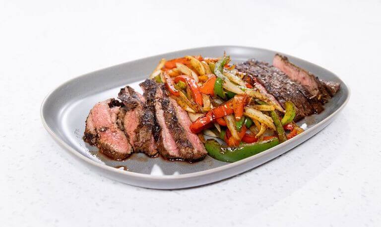 Sliced cuts of beef fanned on a platter with sauteed slices of onion and green and red peppers. 