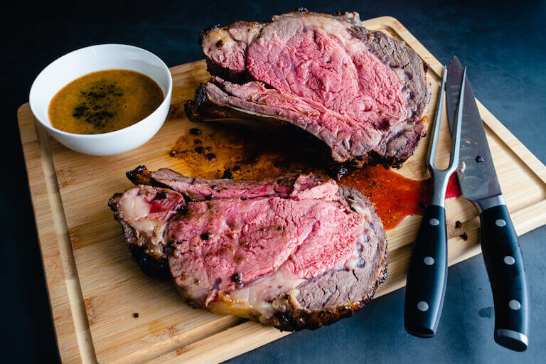 Two medium-rare slices of fat-marbled prime rib roast lay on a cutting board with a meat fork and carving knife. 