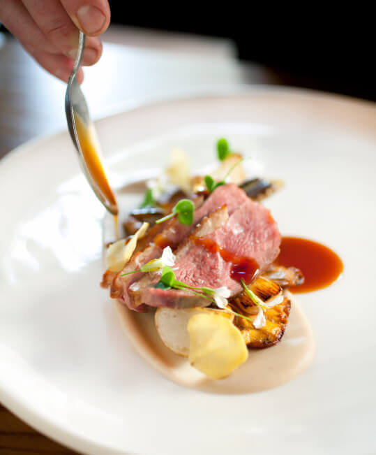 A chef's hand pouring demi-glace sauce with a spoon over a gourmet restaurant plate of duck breast medallions and vegetables