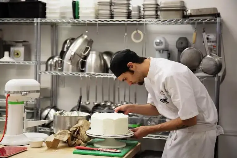 A culinary student in a white uniform and black cap meticulously frost a round cake on a rotating stand in a professional kitchen. Behind, metal shelving is stacked with commercial-grade mixing bowls, pans, and kitchen equipment.
