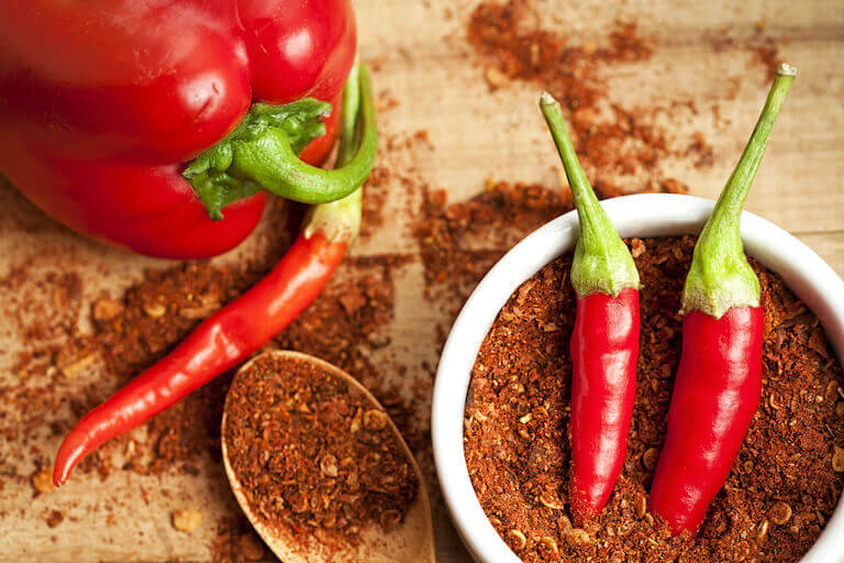 Bowl of ground peppers with chili peppers on top and spices sprinkled on the table around with a whole bell pepper on the side. 