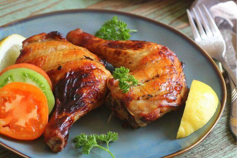 Cooked chicken leg quarters on a blue plate surrounded by red and green tomatoes, lemon slices, and parsley.