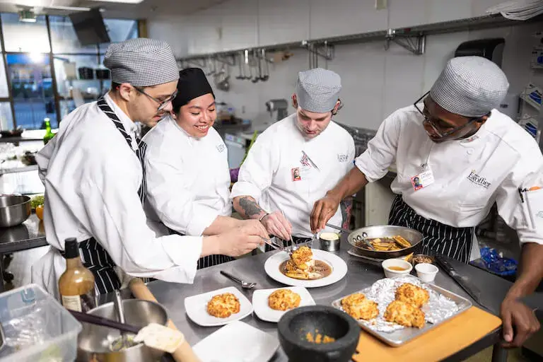 Four culinary students wearing white chef coats and hats gather around a stainless steel prep table in a commercial kitchen. They are smiling and working together to plate a dessert featuring baked goods and sauce, with additional pastries on plates and a baking sheet nearby.