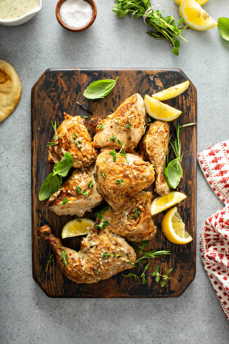Richly roasted chicken pieces like thighs and legs sit on a wooden board decorated with fresh herbs.