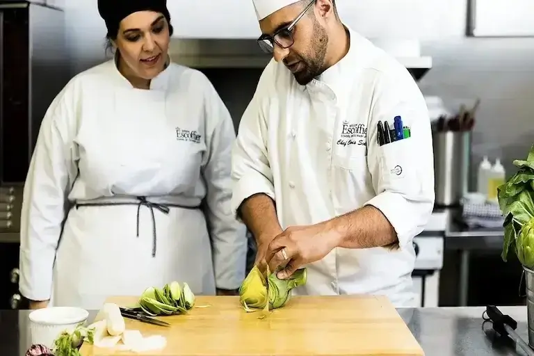 An Escoffier Chef Instructor slices an artichoke on a wooden board while a student observes attentively in the kitchen