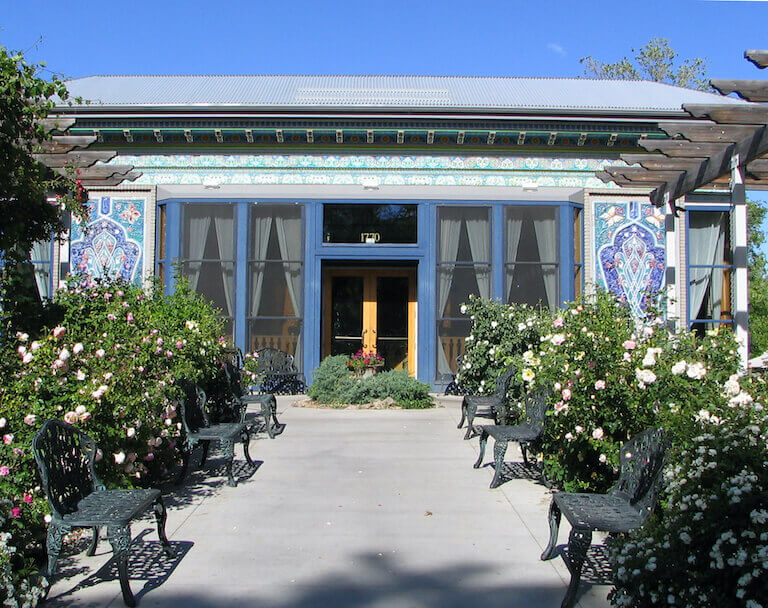 Blooming rose bushes and wrought iron benches line a walkway leading up to the entrance to a low building, flanked by large windows and ornate ceramic tiles.