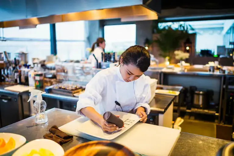 Chef in white coat carefully decorating a dish on a white plate in a professional kitchen.