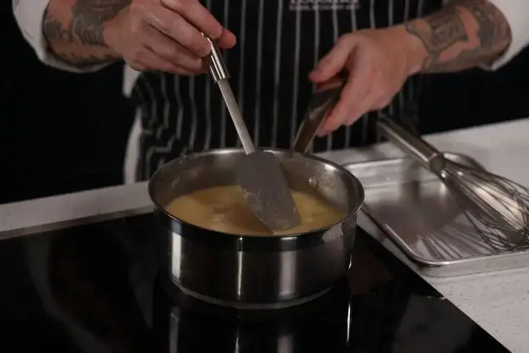 Close-up of a chef with tattooed arms stirring a thick, yellow sauce in a stainless steel pot on an induction cooktop using a grey spatula. A metal tray with a whisk sits on the counter nearby.