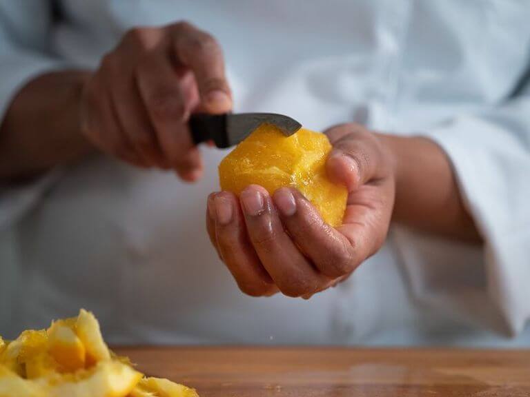 One hand cradles an orange that has been peeled, and the other holds a paring knife and is about to make a cut into the side of the orange.