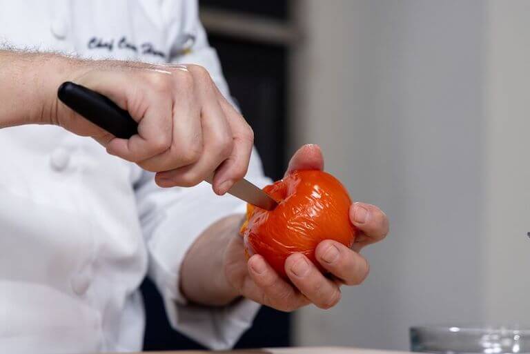 One hand holds a tomato while another holds a paring knife, using the tip of the knife to cut into the top of the tomato in preparation for peeling and deseeding it.