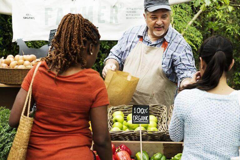 A farmer vendor bags up produce for two women at a farmer’s market stall.
