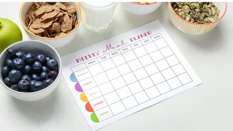 A one-sheet meal planner is shown on a white table and is surrounded by bowls of fresh foods, like blueberries and pumpkin seeds.