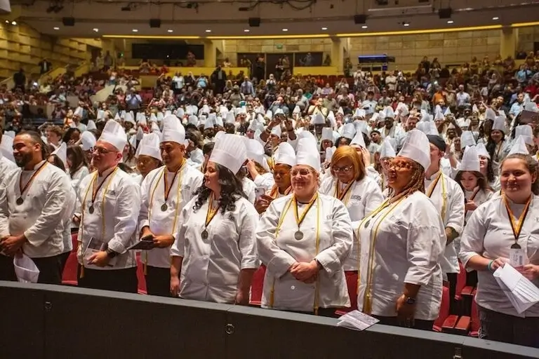 Dozens of people wearing Escoffier-branded chef’s jackets, pleated toques, and medals stand in front of their seats in a large theater during a commencement ceremony, while other attendees look on from rows further back in the theater.