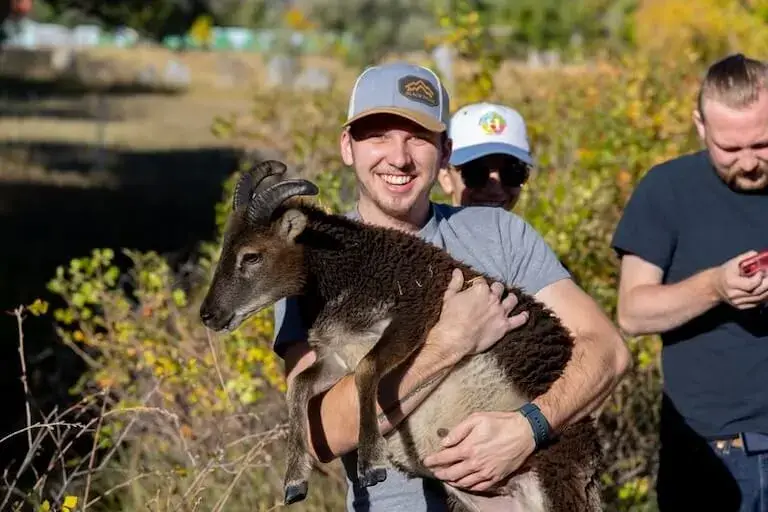 A person wearing a baseball cap and a gray t-shirt stands in a field smiling and holding a goat, with a few other people standing and smiling in the background.