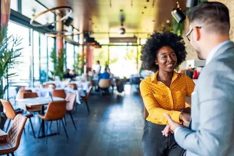 A person with curly hair and a yellow polka-dot shirt smiles and speaks with another person wearing glasses and a suit in a modern restaurant dining room.