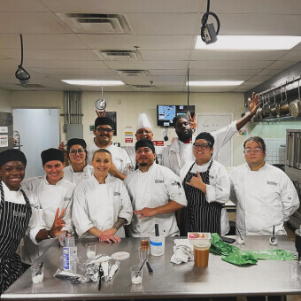 A group of Escoffier culinary arts students with their Chef Instructor smiling in front of a metal table in their professional kitchen classroom