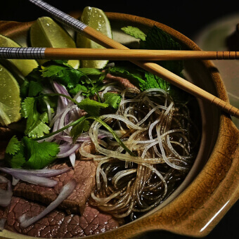 A clear noodle bowl with beef, red onion, cilantro, and lime wedges made by an Escoffier student