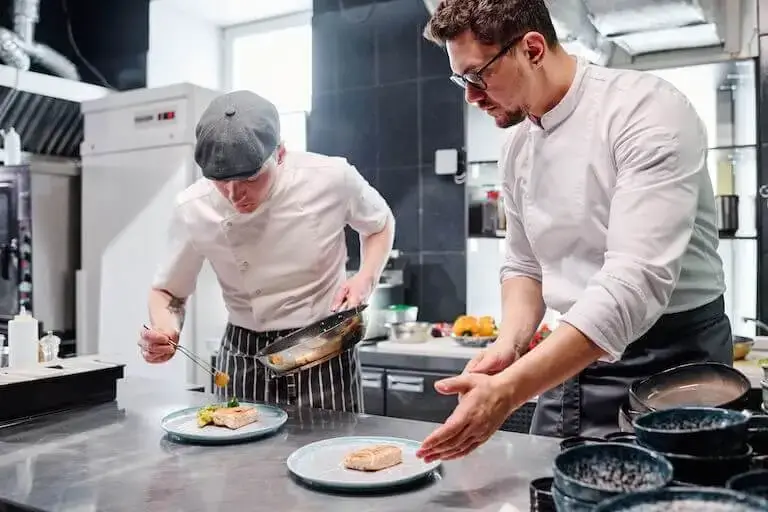 Side view of two chefs in white uniforms carefully plating cooked fish using tweezers and pans on a stainless steel prep station in a modern kitchen.