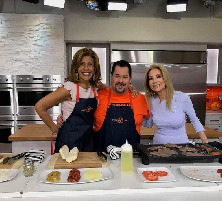 Geno Vento stands between Hoda Kotb and Kathie Lee Gifford, the hosts of NBC’s Today at a demo kitchen counter with thinly sliced steak, sliced cheese, tomato slices, and a squeeze bottle of oil.
