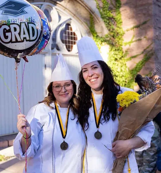 Two smiling Escoffier graduates in white chef uniforms and hats, wearing medals, holding balloons and flowers at a graduation ceremony