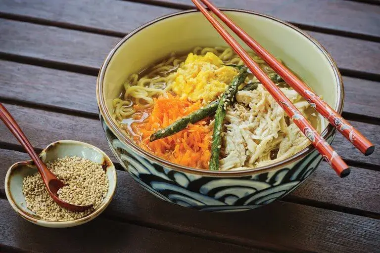 Close-up of a bowl of ginger chicken ramen, featuring sliced chicken, fresh greens, soft-boiled egg, and ramen noodles in a flavorful broth, garnished with sesame seeds.