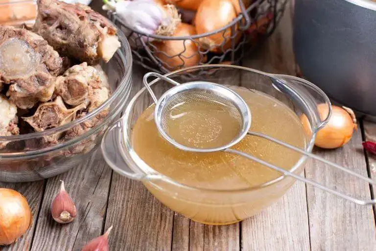 Glass bowl of bone broth with a stainless steel skimmer resting over the bowl.