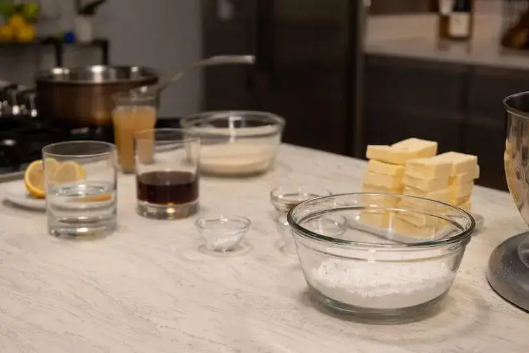 Kitchen counter setup for baking with various ingredients measured in clear glass bowls and glasses. Ingredients include flour, sugar, water, lemon, butter blocks, and vanilla extract, all arranged on a marble countertop.