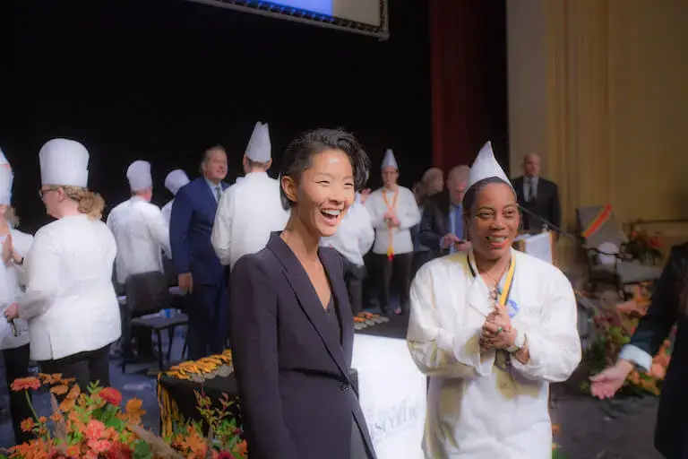 Kristen Kish stands with a graduate at a culinary school graduation. Both are looking at something off-camera and smiling. 