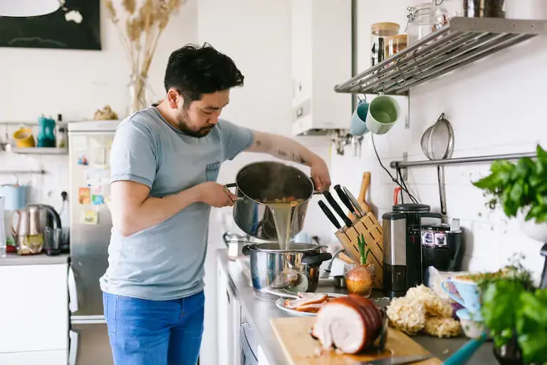 Person in home kitchen straining broth for ramen surrounded by other fresh ingredients on the counter.