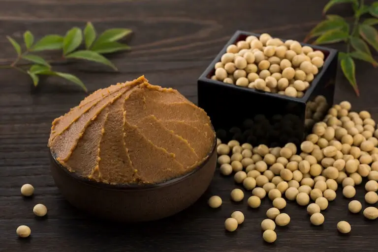 Wooden bowl filled with miso and a dish of soybeans on the table behind it.