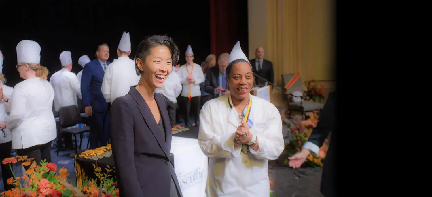 Kristen Kish stands with an Escoffier graduate at the graduation. Both are looking at something off-camera and smiling.
