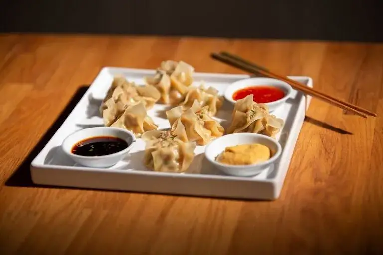 A plate of Siu Mai dumplings served with three dipping sauces on a wooden table.