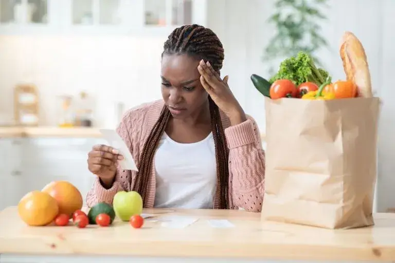A young person sitting in a kitchen surrounded by groceries, scowling while reviewing their grocery receipt. 