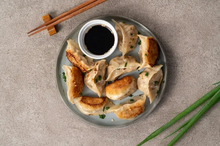 Close-up of pan-fried dumplings with golden edges and soy sauce for dipping, with green onions and chopsticks resting nearby on a stone table.