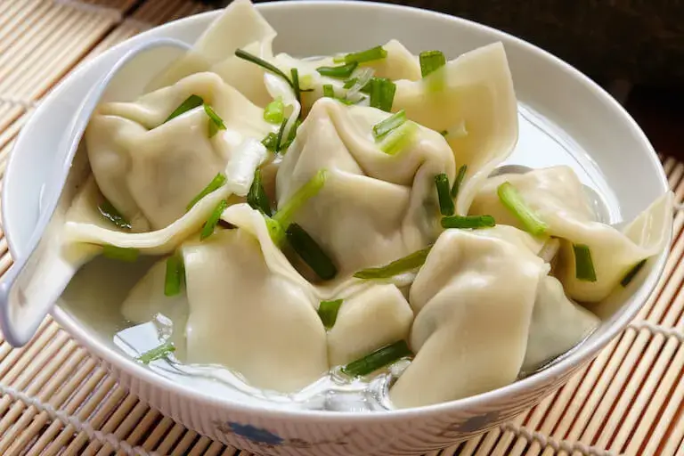 Close-up view of steamed or boiled wonton dumplings served in a shallow white dish with a soup spoon.