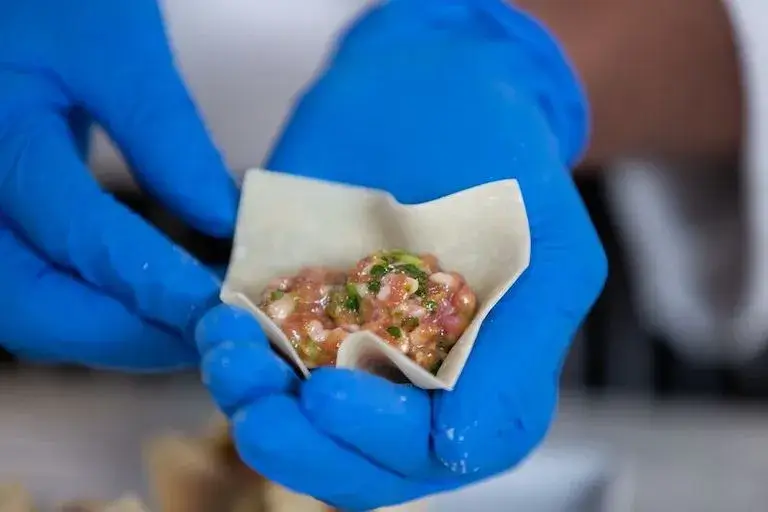 Hands in blue gloves folding a Siu Mai dumpling wrapper filled with a green herb and meat mixture.