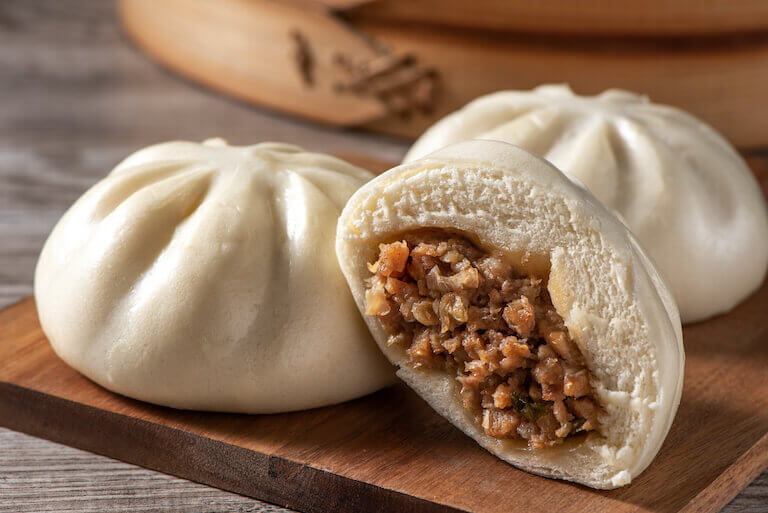 Steamed baozi filled with savory meat, partially open to reveal the filling, resting on a wooden board.