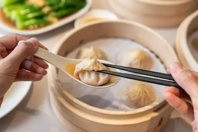 Xiao Long Bao on a spoon, showing the rich, juicy filling and broth, over a bamboo steamer.