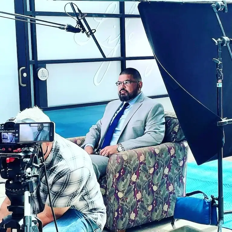 Mike Carrillo wears a suit and tie and sits in an armchair surrounded by film equipment in preparation for an interview.
