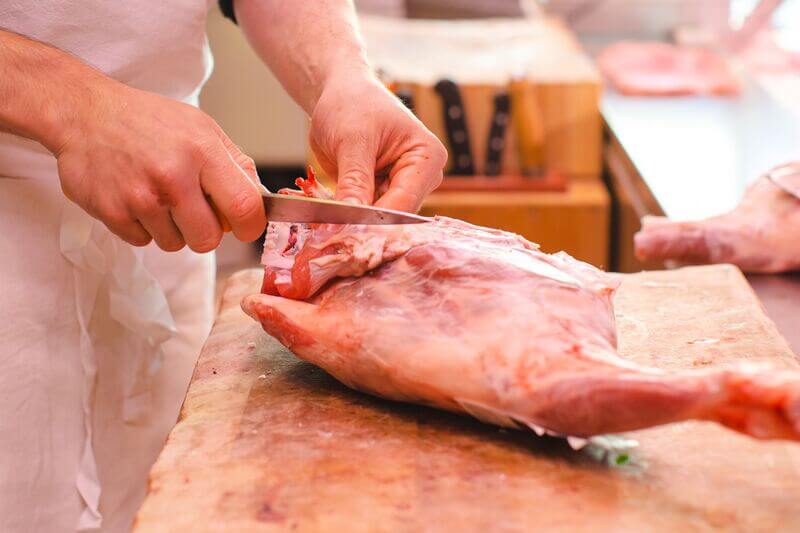 A butcher in a white apron uses a sharp knife to trim and break down a large cut of raw meat on a wooden cutting board.