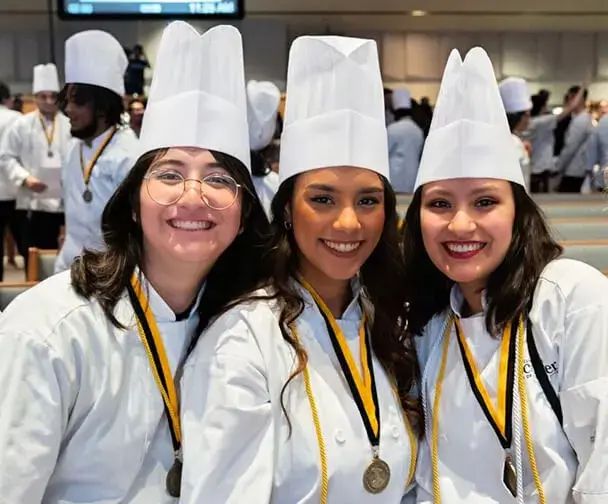 Three Escoffier graduates in chef hats and medals smile at culinary school graduation ceremony