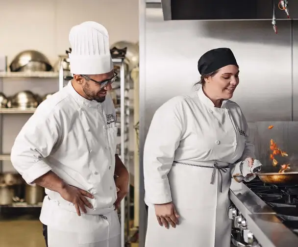 Chef instructor oversees Escoffier culinary student sautéing ingredients over open flame in kitchen