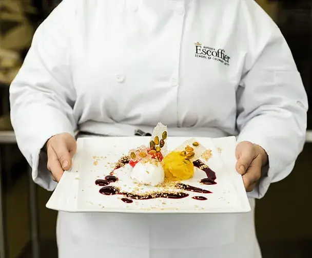 Escoffier culinary student presents plated gourmet dessert with fruit, sauces, and garnishes at Escoffier