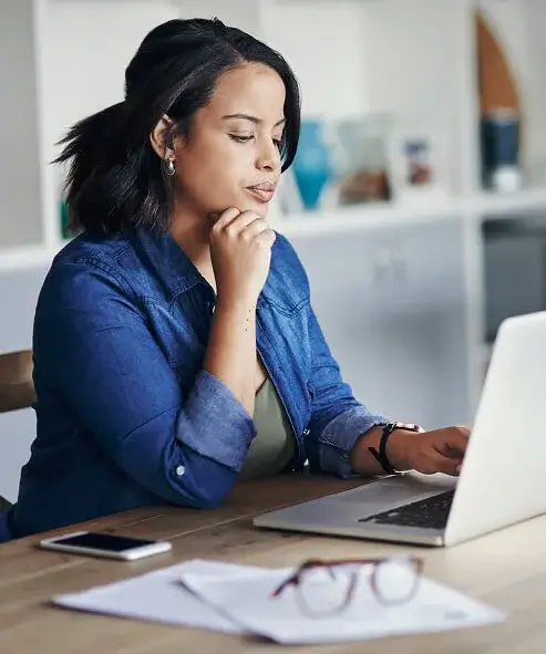 Woman in denim shirt finishing an enrollment application on laptop at home office desk with phone and glasses