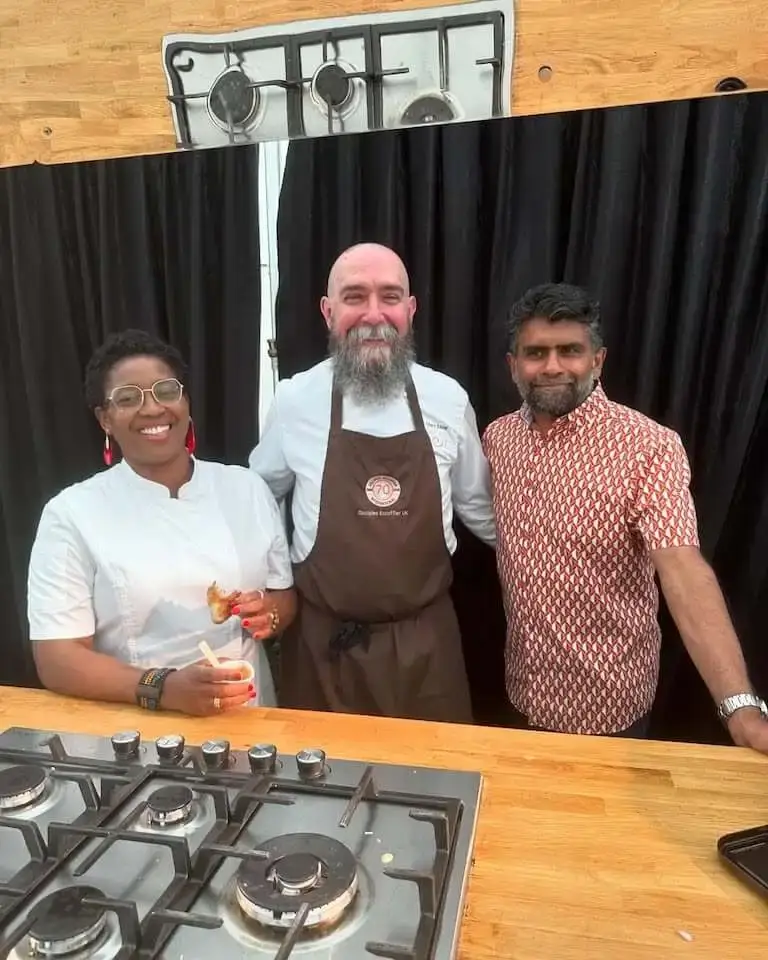 Three people standing together in front of a cooking station, Chef Marc Mole in a brown apron between two other chefs, one holding a small cup of food.